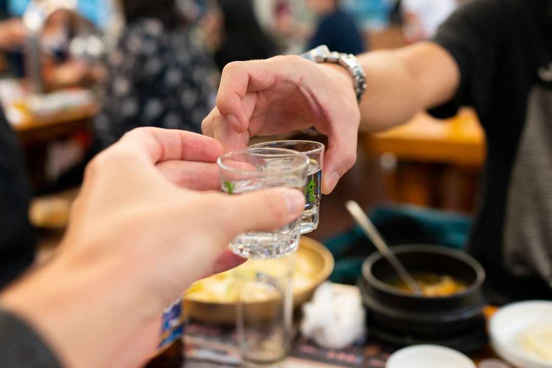 Friends toasting with glasses at a Korean dining table at night with anju food plates, bowls of rice and banchan side dishes, and beverages in a social gathering
